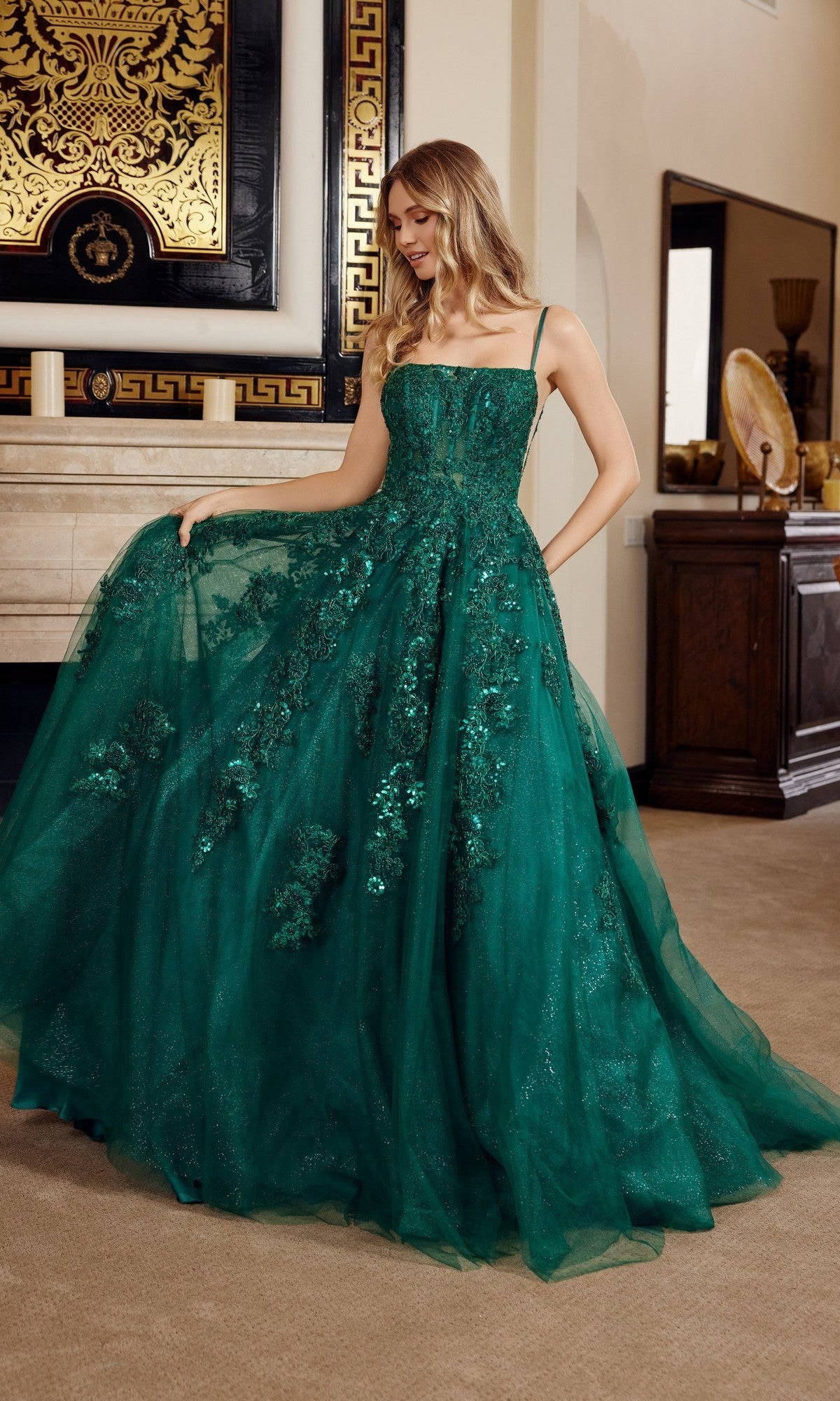 A woman wearing the Nox Anabel H1880 long prom dress—an emerald green, floor-length gown with a sleeveless sequin corset bodice—stands indoors, elegantly holding out part of her dress.